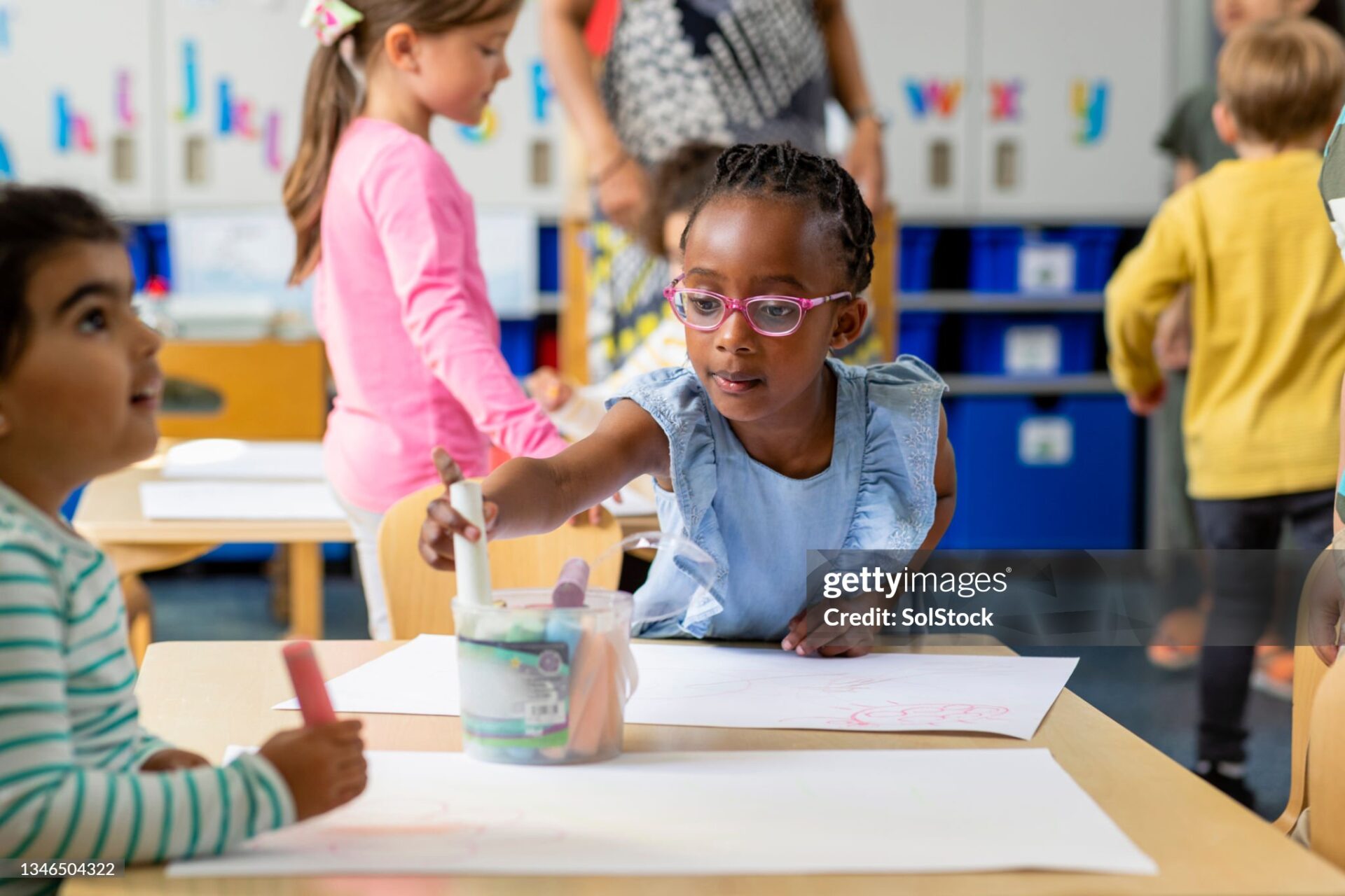 Primary school students sitting in an art classroom drawing the North East of England. The focus is on a girl reaching for a stick of chalk.