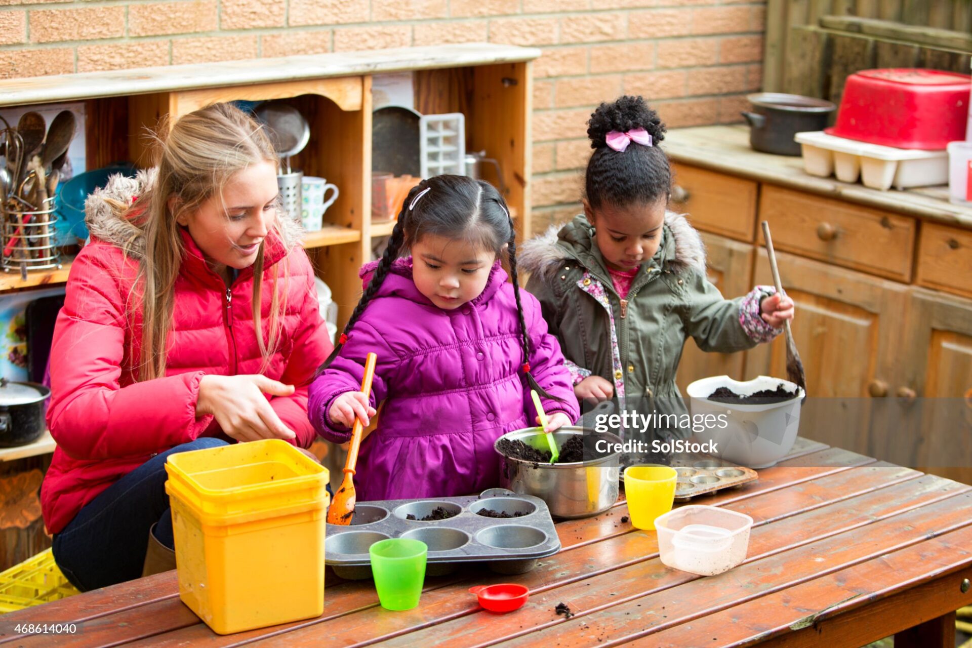 A horizontal image of two children outdoors using a big mental pan to make mud pies, while a young woman supervises. The children are both wrapped up warm in coats and are contently playing