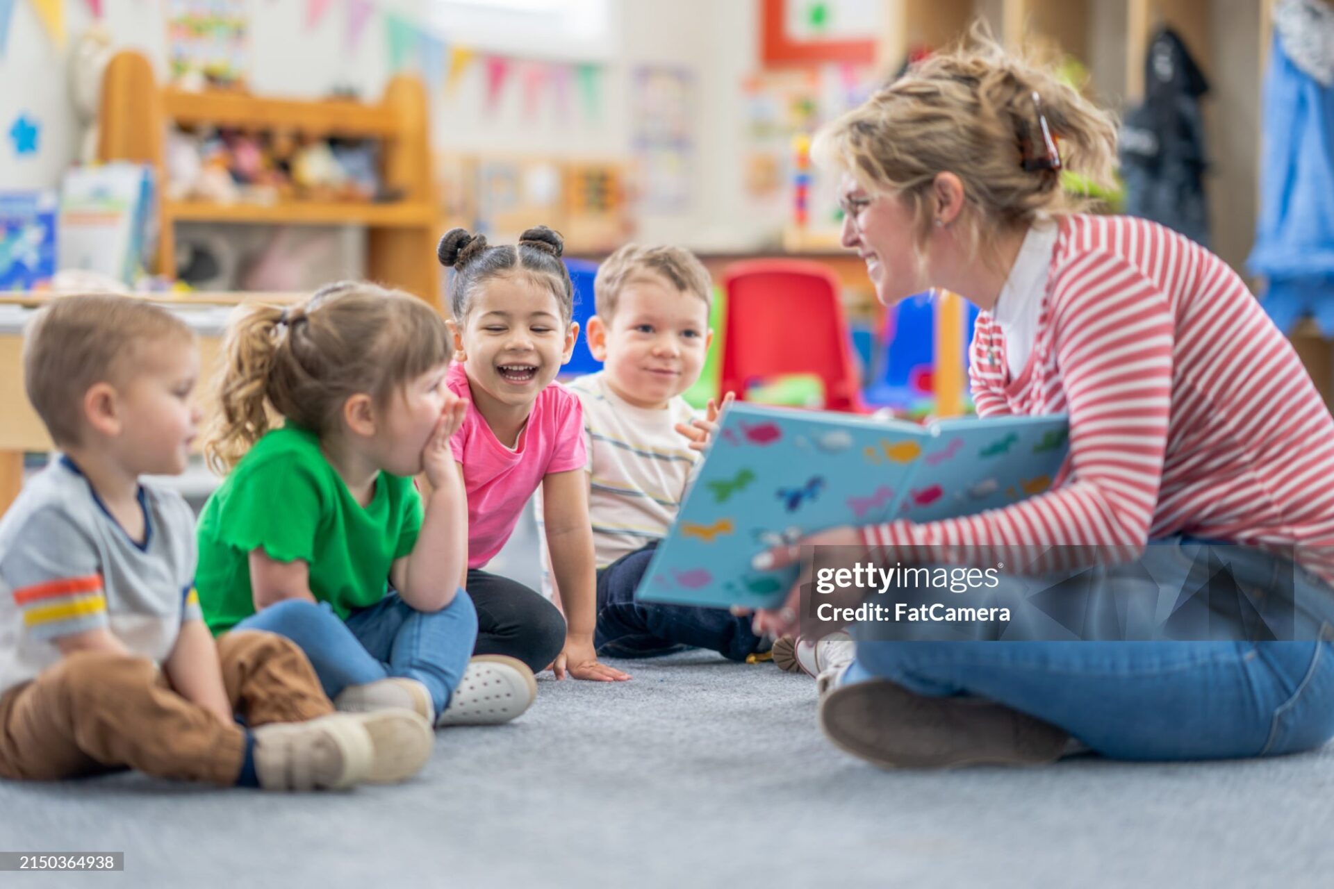 A preschool teacher sits on the floor of her classroom with a small group of students as she reads them a book.  The children are each dressed casually and are focused on the story.